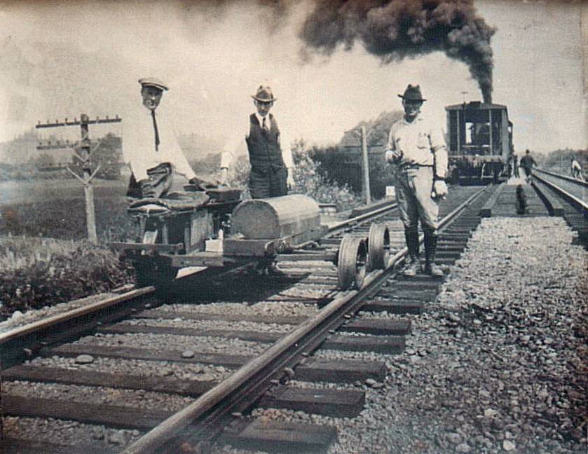 3 well dressed men displaying a Mudge Adams motorcar with a steam locomotive in background